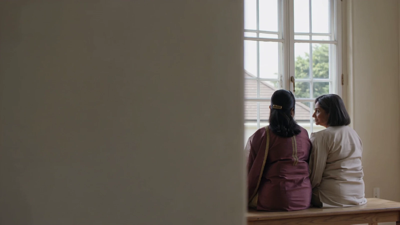 A mother and her adult daughter sit together beside a sunlit window